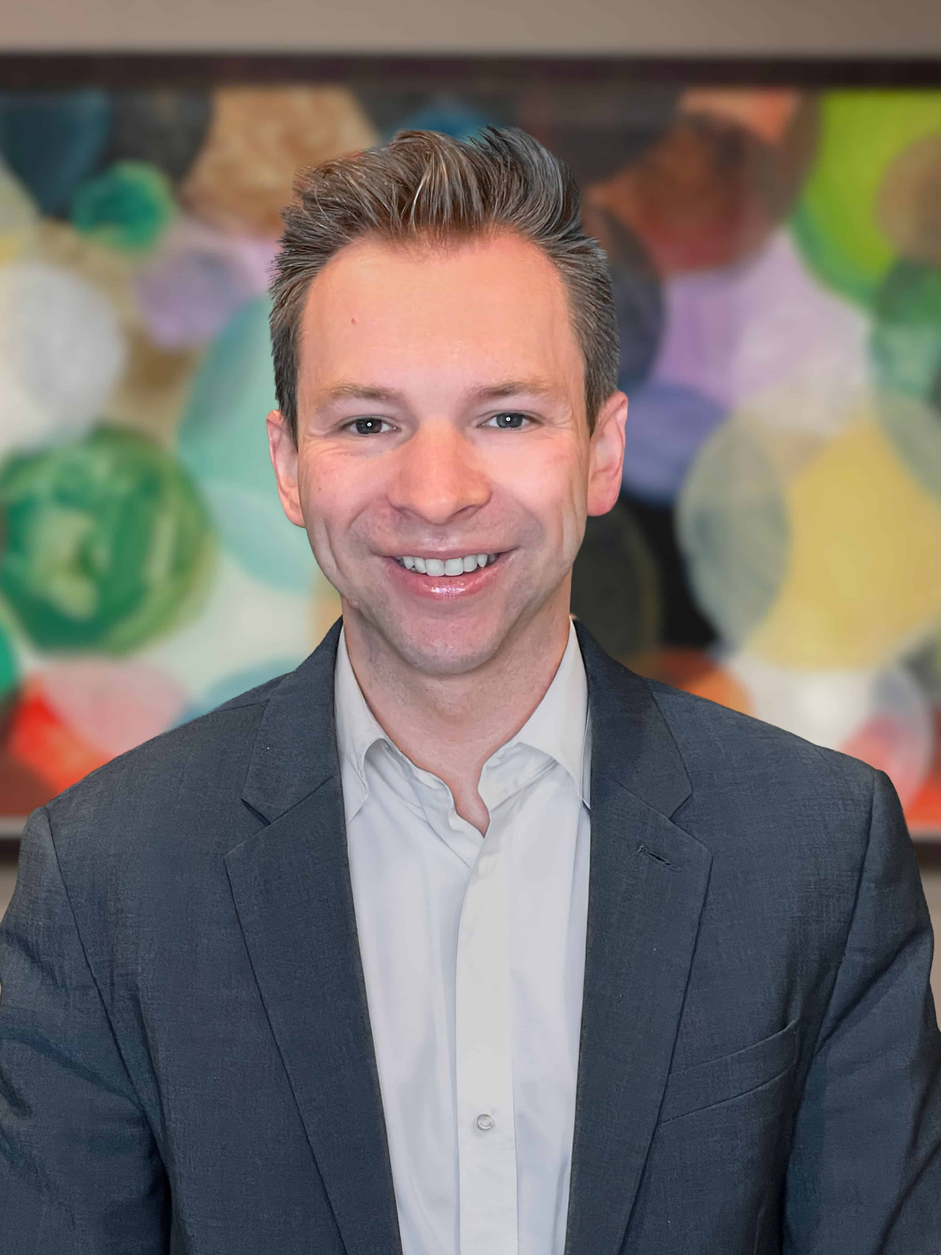 Head-and-shoulders portrait of a smiling man in a dark suit, light shirt, with a colorful abstract mural background