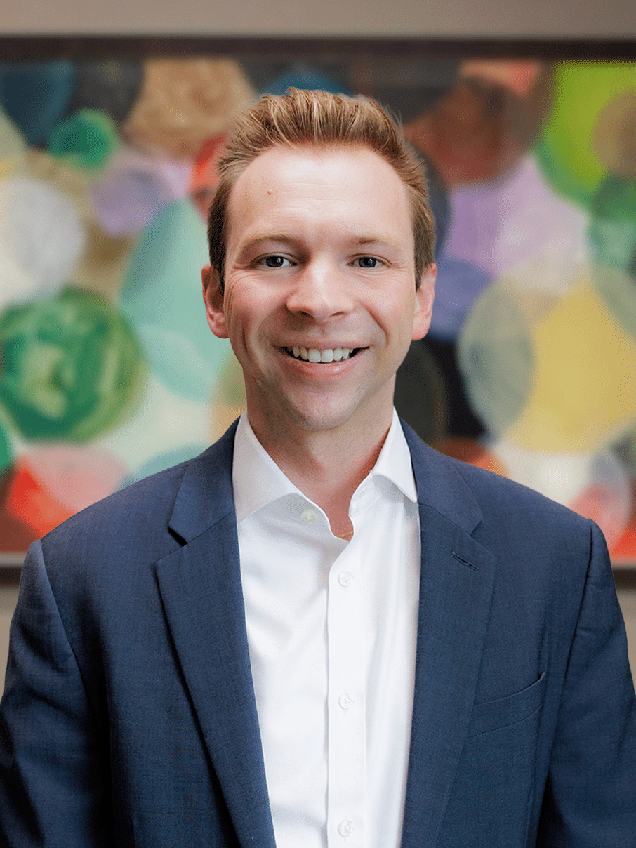 Smiling man in a navy blazer and white shirt posing for a professional portrait with a colorful abstract background.